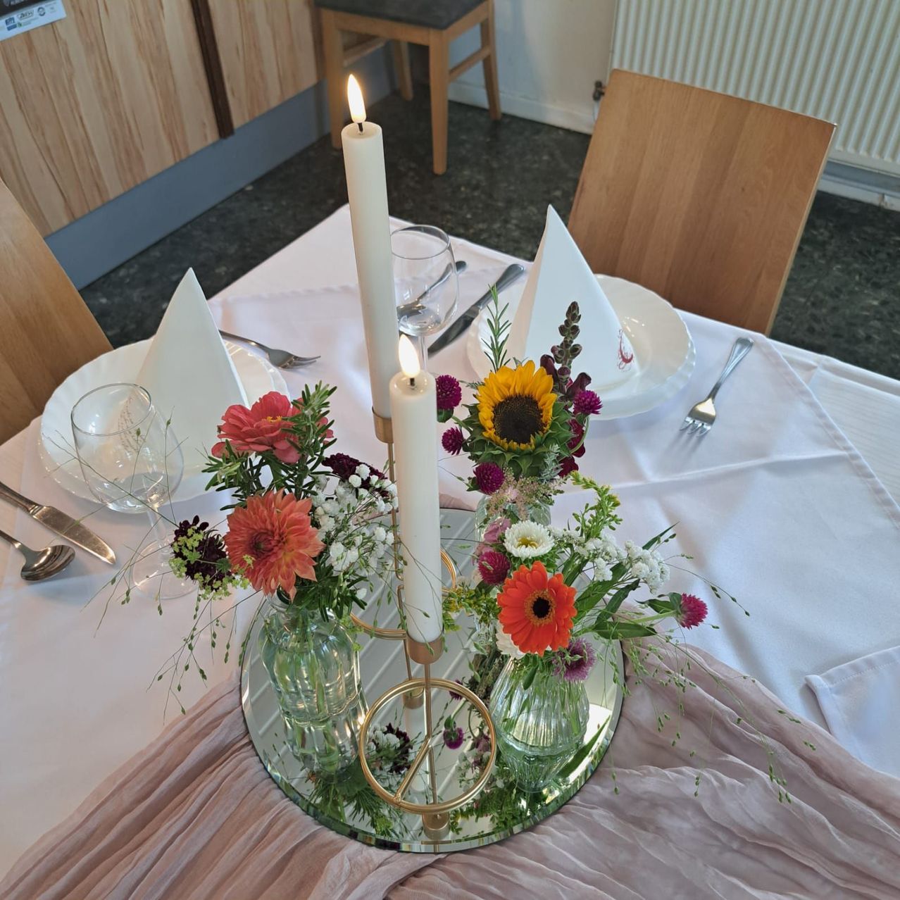 Ein Esstisch ist mit weißen Tellern, Gläsern und Besteck gedeckt. Ein Blumen- und Kerzen-Zentralstück steht auf dem Tisch.