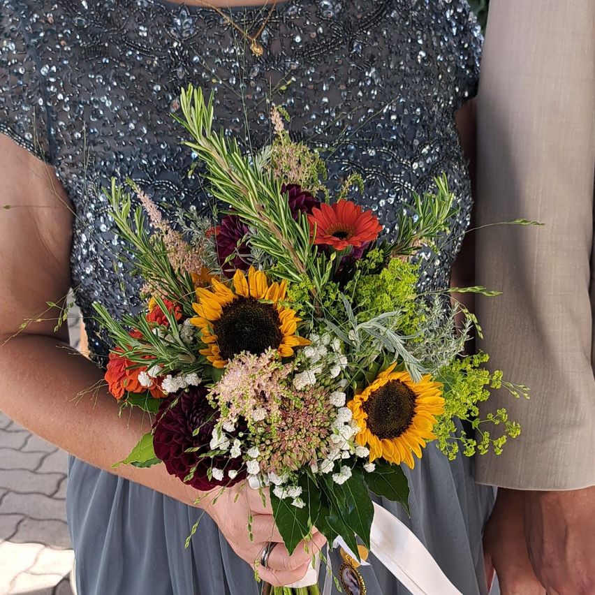 Eine Frau hält einen farbenfrohen Strauß aus Sonnenblumen, roten und weißen Blumen und Grün. Sie trägt ein Paillettenkleid.