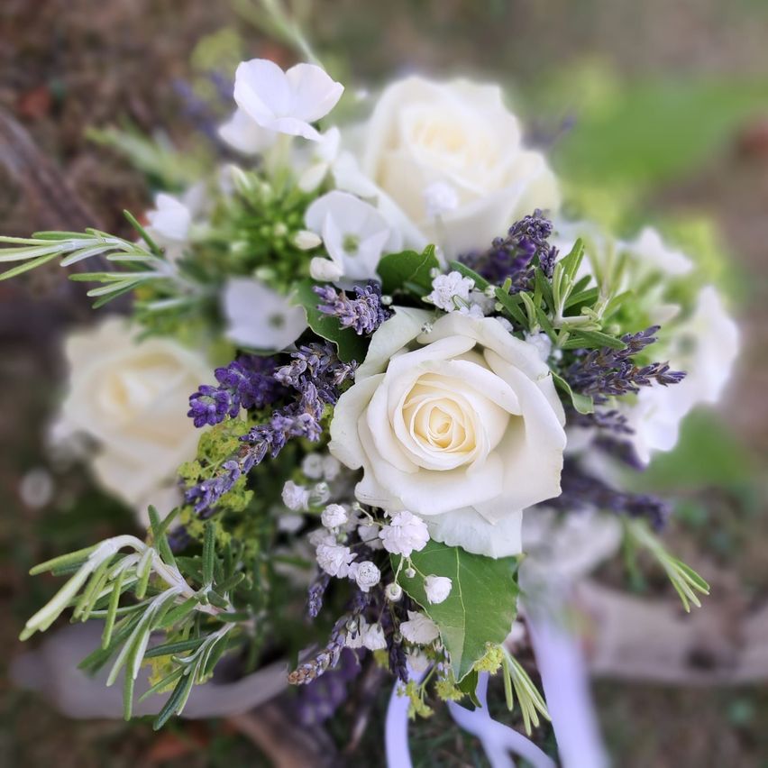 Ein Nahaufnahme eines Brautstraußes mit weißen Rosen, Lavendel und grünen Blättern. Die Rosen sind in einer kreisförmigen Form angeordnet, mit einigen Blättern verstreut.