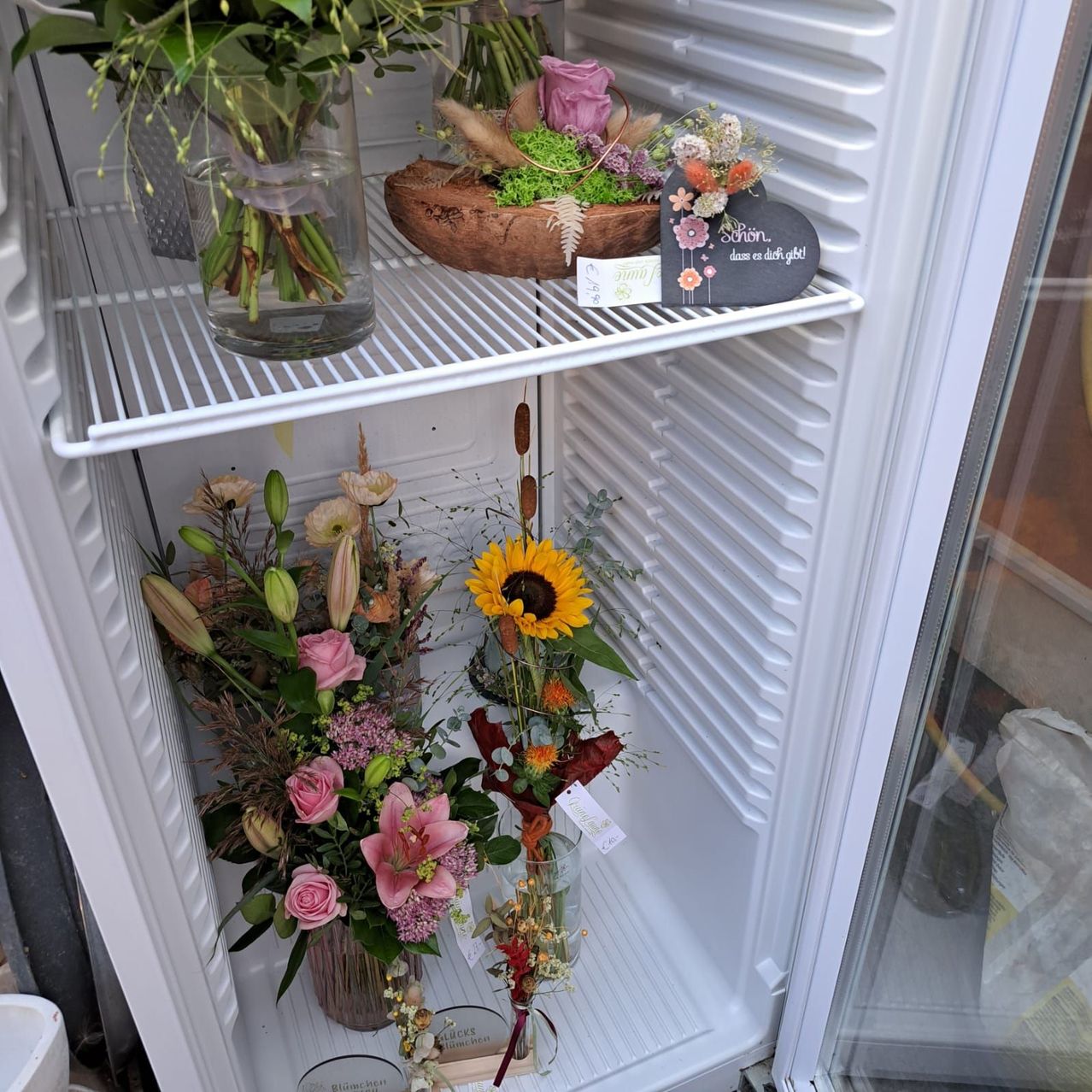 A refrigerator with three shelves, each holding different floral arrangements. The top shelf has a vase with green leaves, the middle has a sunflower, and the bottom has a mix of pink and white flowers.