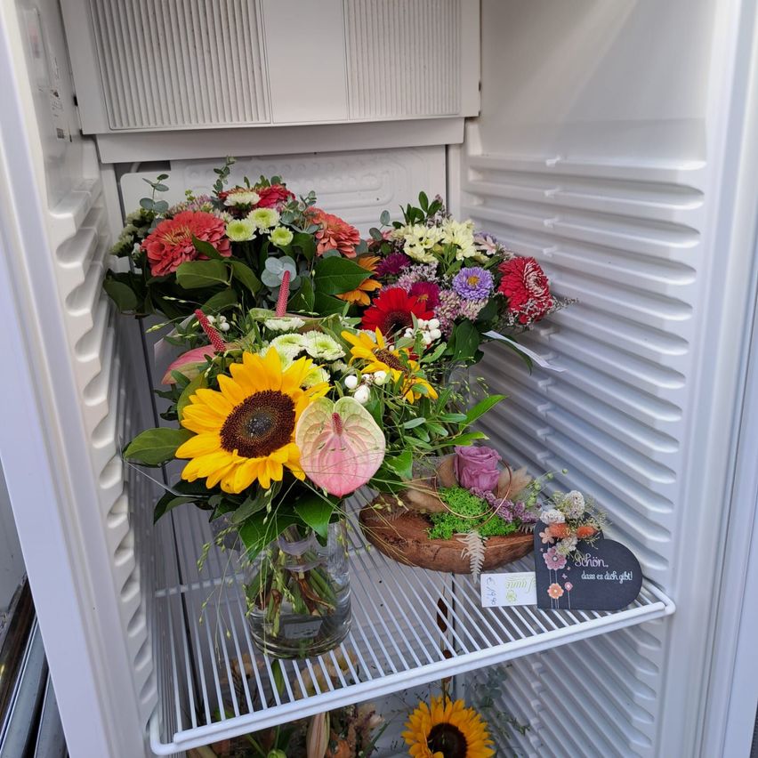 A refrigerator door displays an array of vibrant flowers, including sunflowers, roses, and daisies, arranged in vases and pots on its shelves. A chalkboard sign reads 'Enjoy the day!' in German.