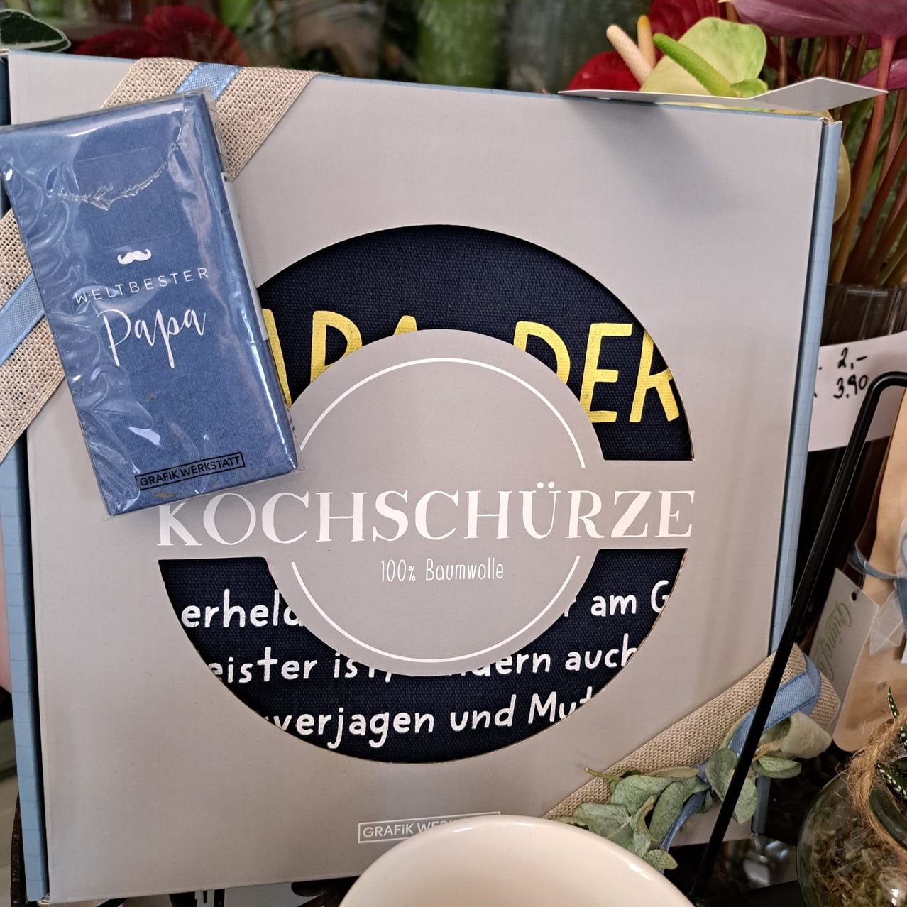 Ein Geschenkbox für Väter mit einer blau-weißen Schürze. Es steht 'KOCHSCHURZ' und '100% Baumwolle'. Ein Geschenkanhänger mit 'Weltbester Papa' ist angebracht.