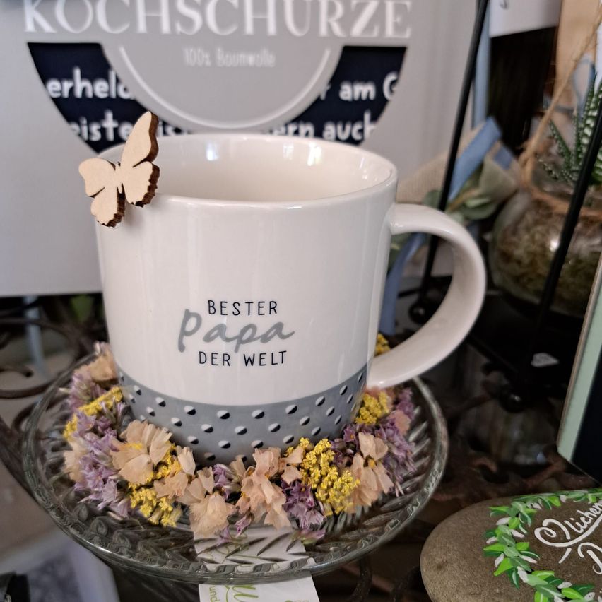 Eine weisse Tasse mit grauen Punkten und dem Text 'Bester Papa der Welt' steht auf einer Glasplatte mit Blumen. Ein Schmetterling sitzt auf der Tasse.