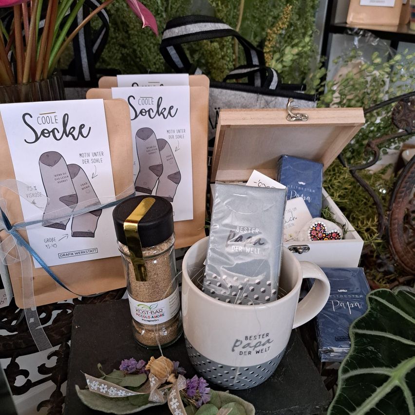 Ein Tisch zeigt eine weiße Kaffeetasse, einen Beutel mit Gewürzen, ein Geschenksbox und eine Packung Socken. Es gibt auch Pflanzen und Blumen auf dem Tisch.