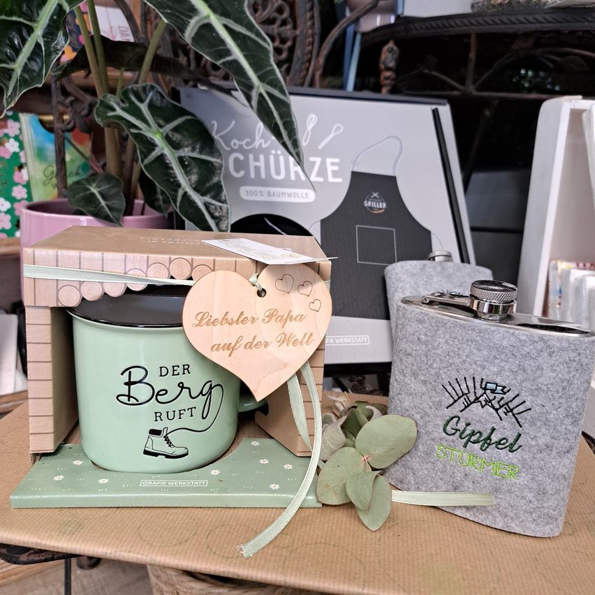 Ein Geschenkset mit einer grünen Tasse, die mit 'Der Berg Ruft' beschriftet ist, einer grauen Flasche mit der Aufschrift 'Gipfelstürmer' und einem herzförmigen Anhänger. Die Tasse ist in einer Schachtel mit einer Schleife, und das Set steht auf einem Tisch mit einer Pflanze im Hintergrund.