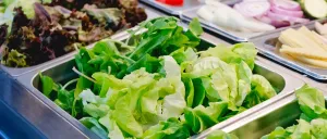Mehrere Tabletts mit frischen Salaten, einschließlich Salat, sind ausgestellt. Andere Gemüsesorten wie Eier, Tomaten und Zwiebeln sind ebenfalls vorhanden.