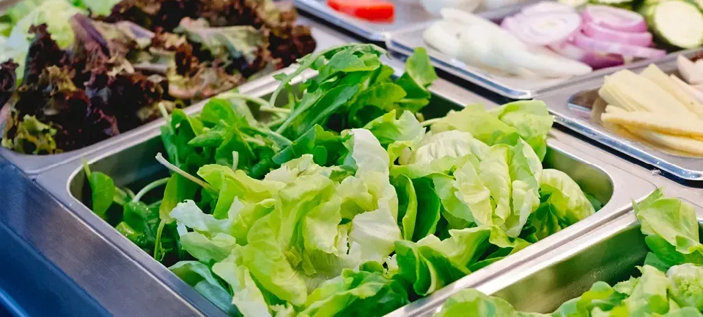 Mehrere Tabletts mit frischen Salaten, einschließlich Salat, sind ausgestellt. Andere Gemüsesorten wie Eier, Tomaten und Zwiebeln sind ebenfalls vorhanden.