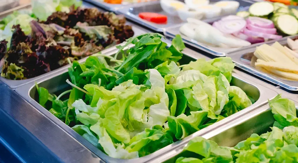 Mehrere Tabletts mit frischen Salaten, einschließlich Salat, sind ausgestellt. Andere Gemüsesorten wie Eier, Tomaten und Zwiebeln sind ebenfalls vorhanden.