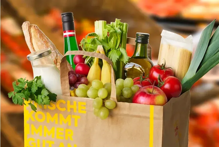 Ein brauner Papierbeutel gefüllt mit Brot, Gemüse, Früchten und Getränken. Der Beutel trägt die Aufschrift 'FRISCHE KOMMT IMMER GUT AN'.