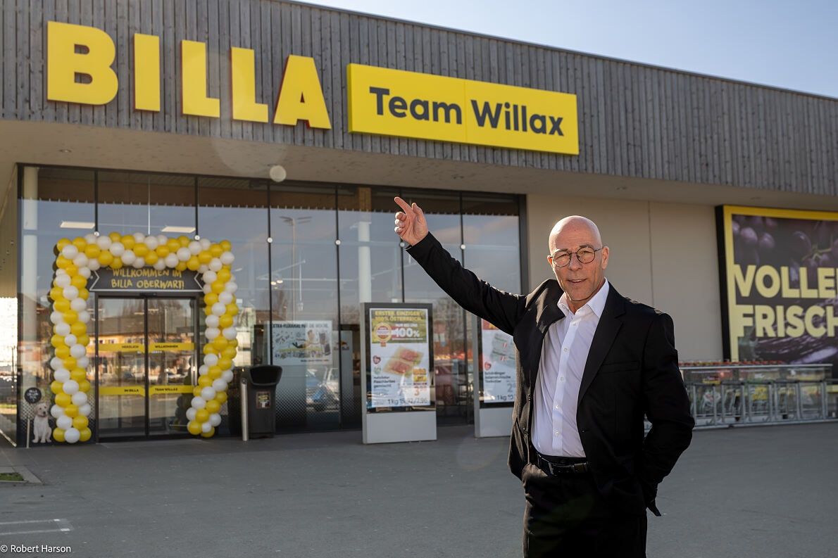 Ein Mann im Anzug steht vor einem Geschäft mit einem gelben Schild, das Team Willax liest. Es gibt Ballons und einen Mülleimer in der Nähe.
