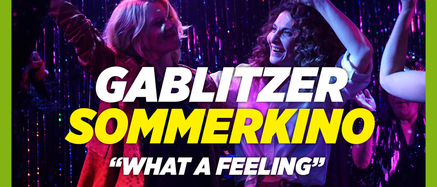 Zwei Frauen lächeln auf der Bühne mit dem Text 'GABLITZER IMMERKIN' und 'What a feeling' darunter. Sie scheinen zu performen oder zu feiern.