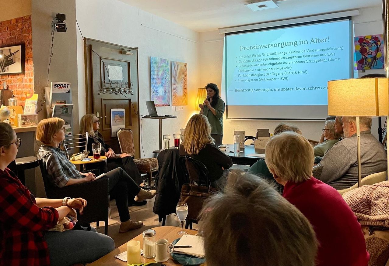 Eine Frau spricht in ein Mikrofon in einem Raum mit einer Projektionsleinwand. Menschen sitzen an Tischen, einige mit Gläsern und Notizen. Die Leinwand zeigt Text über Proteinaufnahme im Alter.