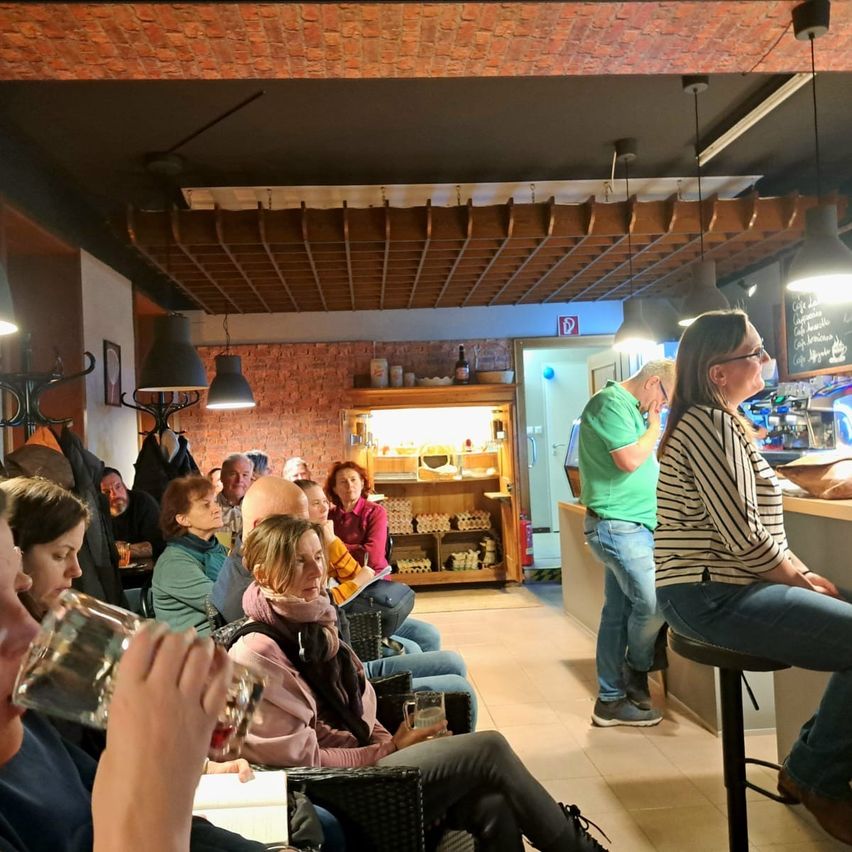 Eine Indoor-Versammlung in einem Café, eine Person trinkt aus einem Glas, eine Frau schaut auf eine Speisekarte und ein Mann steht hinter der Theke.