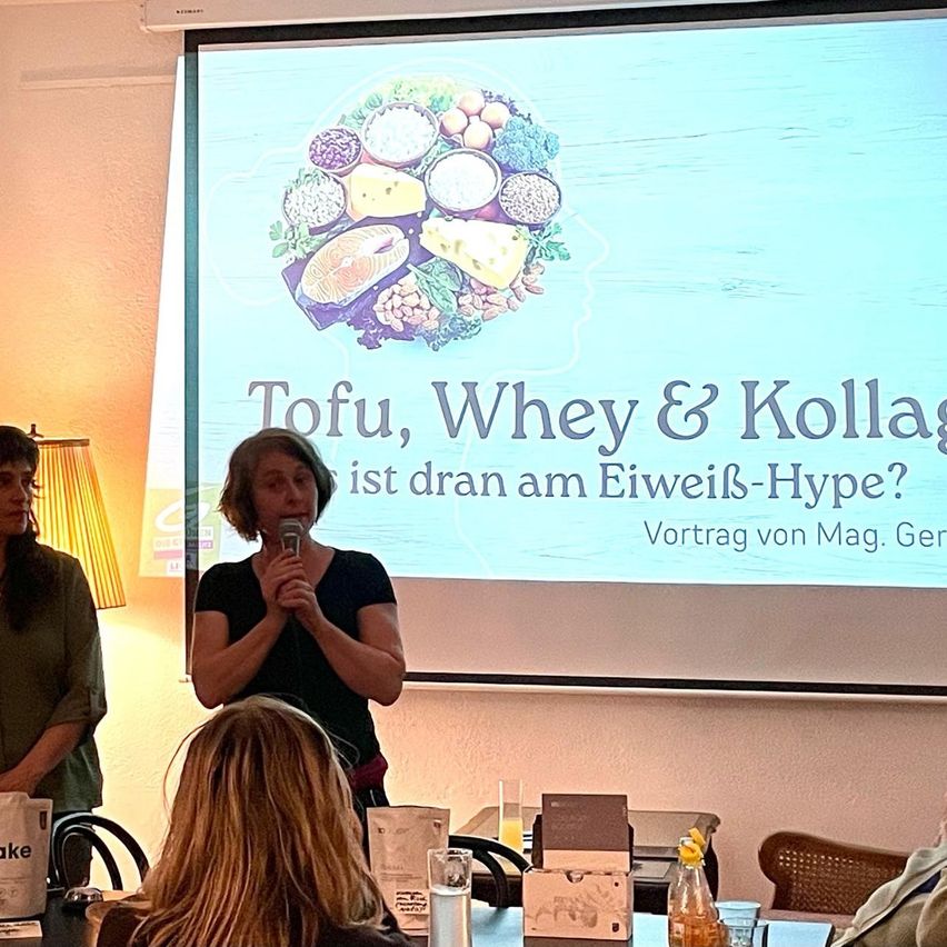 Eine Frau spricht in ein Mikrofon in einem Raum. Eine Leinwand hinter ihr zeigt eine Präsentation über Tofu, Whey und Kollagen. Mehrere Personen sitzen an einem Tisch und hören zu.