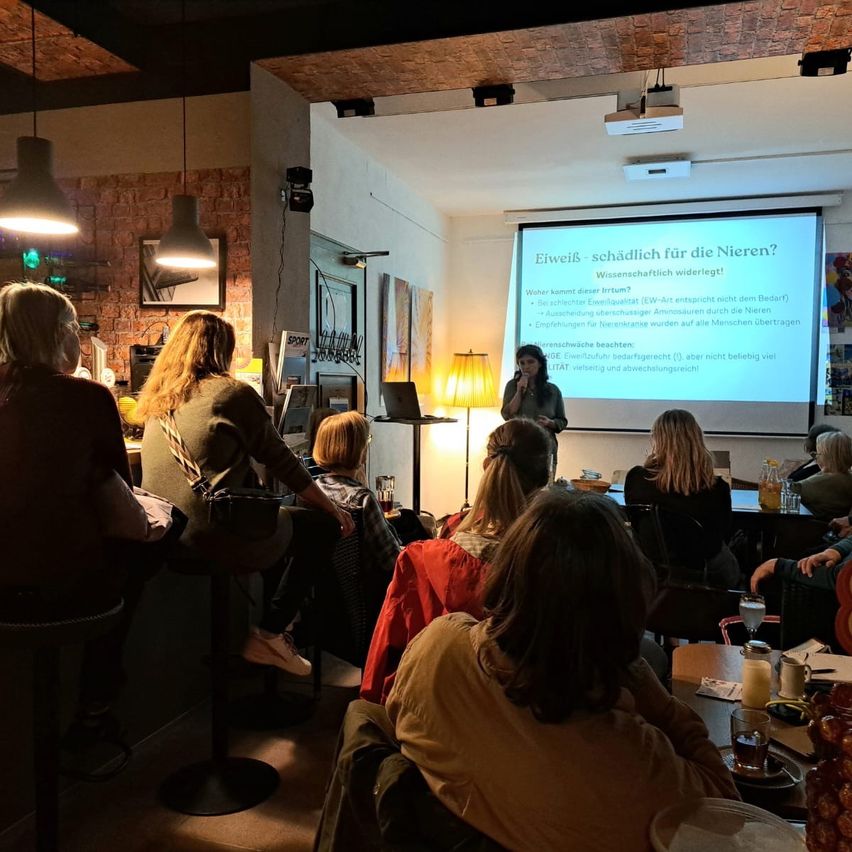 Eine Frau hält eine Präsentation über Nierengesundheit bei einer Versammlung ab, wobei ein Beamer damit verbundene Informationen anzeigt. Die Menschen sitzen um Tische herum und hören aufmerksam zu.