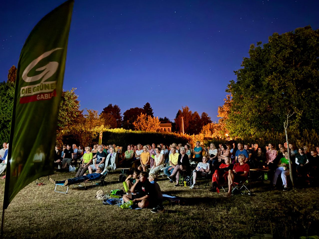 Eine große Gruppe von Menschen versammelt sich bei Nacht für eine Veranstaltung im Freien, sitzt auf Stühlen und Decken unter einem Sternenhimmel. Bäume und ein Gebäude sind im Hintergrund zu sehen.