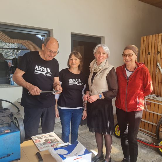 Vier Menschen stehen in einem Raum, lächeln und posieren für ein Foto. Der Mann links hält ein Werkzeug, und eine Frau in der Mitte hält ein Stück Papier. Der Raum hat eine Holzwand und eine Tür. Auf der rechten Seite befindet sich eine Holzkonstruktion mit einem gelben Rad.