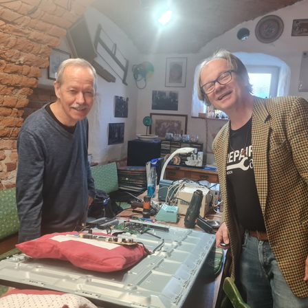 Zwei Männer stehen in einem Raum mit einer Ziegelwand, lächeln und posieren für ein Foto. Ein Tisch vor ihnen hat einen Laptop und andere elektronische Gegenstände. Ein grüner Stuhl und ein Regal mit Gegenständen sind hinter ihnen.