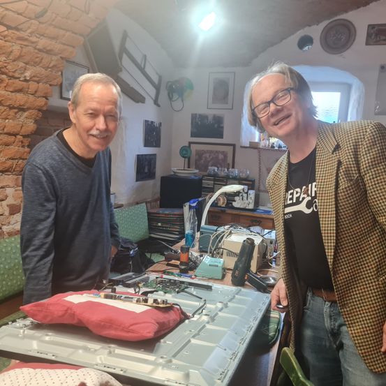 Zwei Männer stehen in einem Raum mit einer Ziegelwand, lächeln und posieren für ein Foto. Ein Tisch vor ihnen hat einen Laptop und andere elektronische Gegenstände. Ein grüner Stuhl und ein Regal mit Gegenständen sind hinter ihnen.