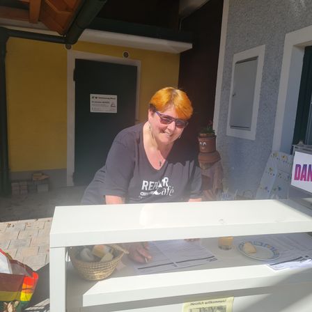 Eine Frau mit orangefarbenen Haaren und einer Brille steht hinter einem Tresen eines kleinen Unternehmens. Sie trägt ein schwarzes Shirt mit dem Wort Rear Cafe.