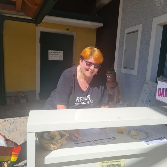 Eine Frau mit orangefarbenen Haaren und einer Brille steht hinter einem Tresen eines kleinen Unternehmens. Sie trägt ein schwarzes Shirt mit dem Wort Rear Cafe.