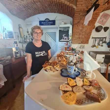 Eine Frau mit Brille und einem schwarzen T-Shirt mit dem Wort Reparatur-Café steht hinter einem Tisch mit verschiedenen Speisen darauf.