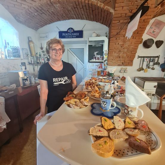 Eine Frau mit Brille und einem schwarzen T-Shirt mit dem Wort Reparatur-Café steht hinter einem Tisch mit verschiedenen Speisen darauf.