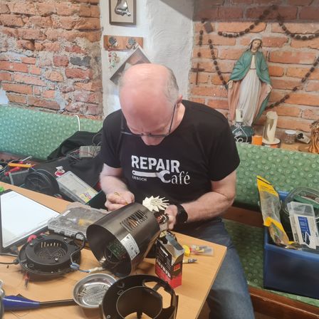 Ein Mann in einem schwarzen T-Shirt mit dem Wort Reparatur darauf repariert ein schwarzes elektrisches Gerät auf einem Holztisch.