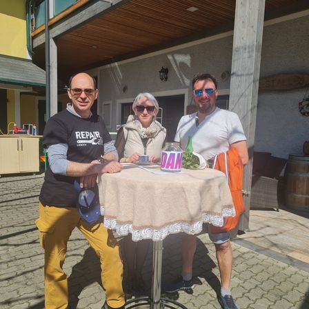Drei Personen stehen um einen Tisch und lächeln für ein Foto. Ein Mann links trägt ein schwarzes T-Shirt mit dem Wort 'REPAIR'. Eine Frau sitzt in der Mitte und lächelt. Ein Mann rechts trägt eine Sonnenbrille und eine orange Weste.