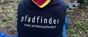 Ein junger Junge sitzt mit dem Rücken zur Kamera in einem Wald und trägt ein blaues T-Shirt mit 'pfadfinder' und 'scout.at/oberpullendorf' darauf.