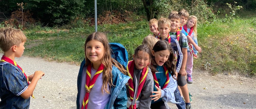 Eine Gruppe von Kindern in Pfadfinderuniformen steht auf einem Kiesweg. Hinter ihnen befindet sich ein Schild, und sie lächeln und posieren für ein Foto.
