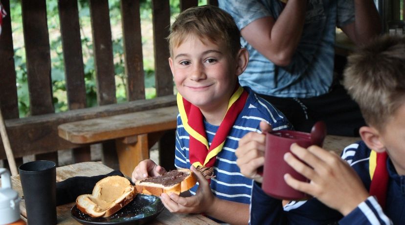 Zwei Jungen in Pfadfinderuniformen essen Sandwiches an einem Picknicktisch. Einer hält einen Becher, während der andere ein halb gegessenes Sandwich hat. Eine Holzbank und eine weitere Person sind im Hintergrund.
