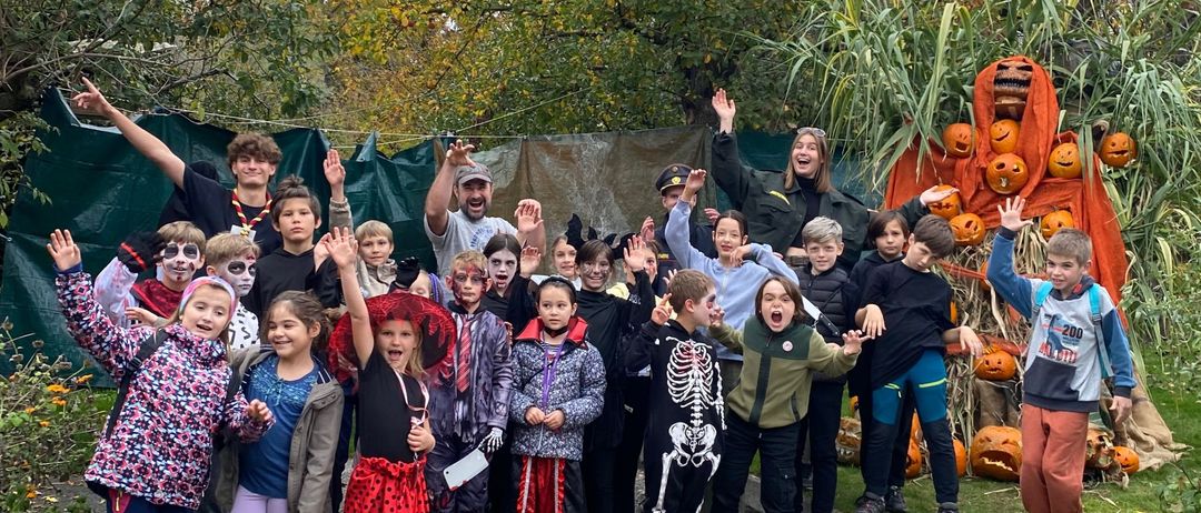 Eine Gruppe von Kindern in Halloween-Kostümen posiert für ein Foto in einem Außenbereich mit Kürbissen und Bäumen im Hintergrund.
