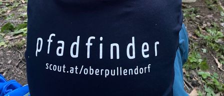 Ein Junge mit dem Wort Pfadfinder auf dem Rücken sitzt auf dem Boden, während andere in einem Wald hinter ihm stehen.