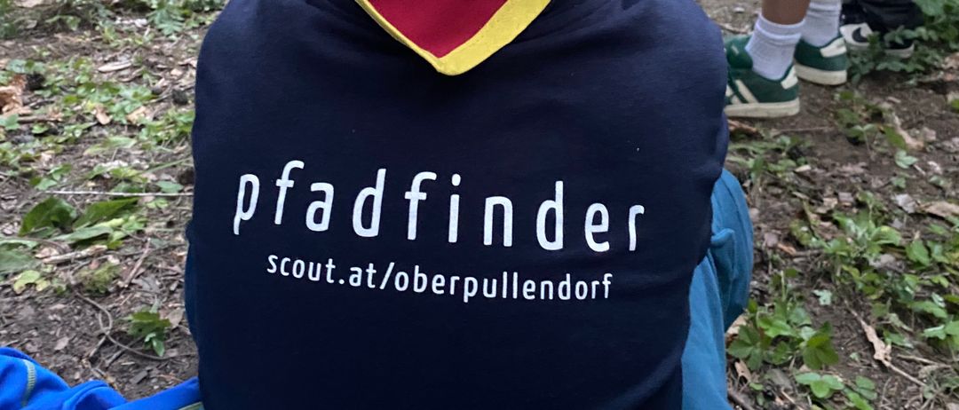 Ein Junge in einem Pfadfinder-Shirt sitzt auf dem Boden, während andere hinter ihm in einem Waldgebiet stehen.