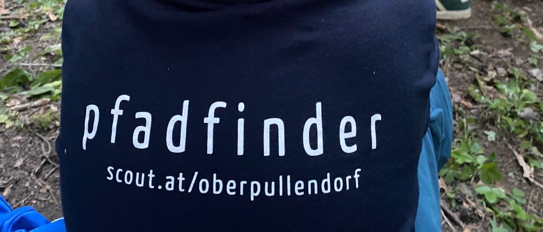 Ein Junge mit einem Pfadfinder-Hemd sitzt draußen, mit anderen Kindern und Erwachsenen um ihn herum. Die Umgebung scheint ein Wald oder ein bewaldetes Gebiet zu sein.