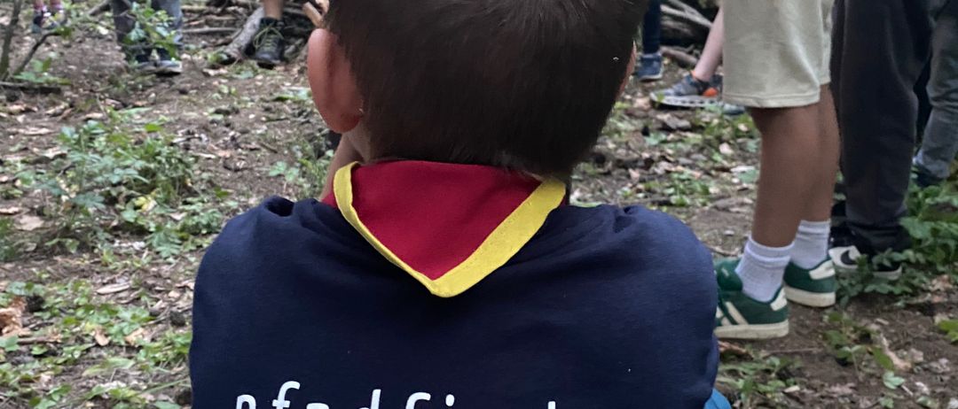 Ein Junge mit einem Pfadfinder-Hemd sitzt in einem Wald, mit einer Gruppe von Leuten um ihn herum, einige stehen und andere sitzen auf einem Baumstamm.