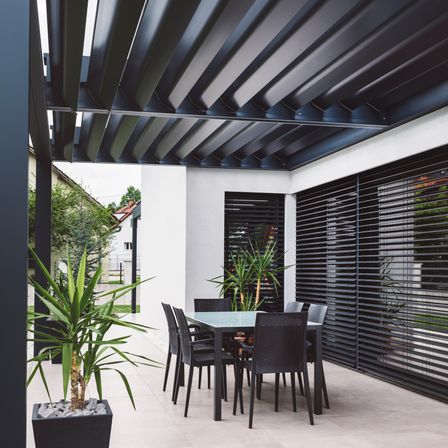 Ein moderner Terrassenbereich mit Tisch, Stühlen und Pflanzen. Schwarze Markisen und Jalousien bieten Schatten und Privatsphäre. Die Terrasse ist von einem Fliesenboden und einer weißen Wand umgeben.