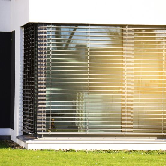 Ein modernes Haus außen mit einem großen Fenster, das von weißen Jalousien bedeckt ist. Das Sonnenlicht scheint durch die Jalousien und wirft einen gelben Schimmer auf das Fenster.