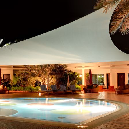 Ein Swimmingpool, umgeben von Liegestühlen und einem Vordach, bei Nacht beleuchtet. Bäume und Pflanzen umgeben das Gebiet.