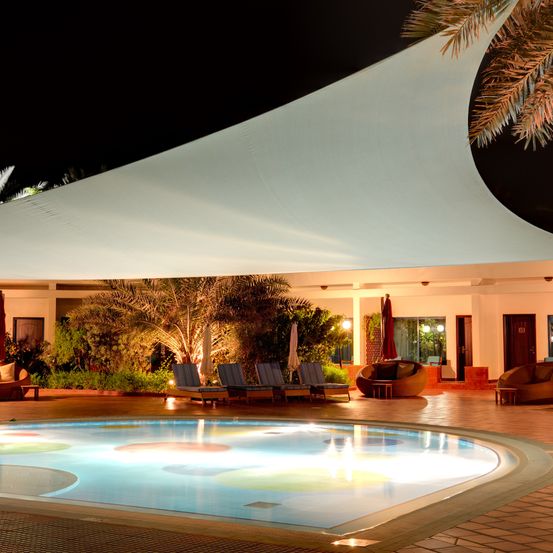 Ein Swimmingpool, umgeben von Liegestühlen und einem Vordach, bei Nacht beleuchtet. Bäume und Pflanzen umgeben das Gebiet.
