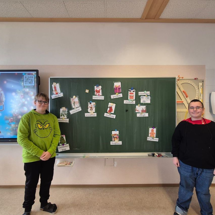 Zwei Jungen stehen neben einer Klassentafel mit weihnachtlichen Karteikarten und einem großen Monitor im Hintergrund.