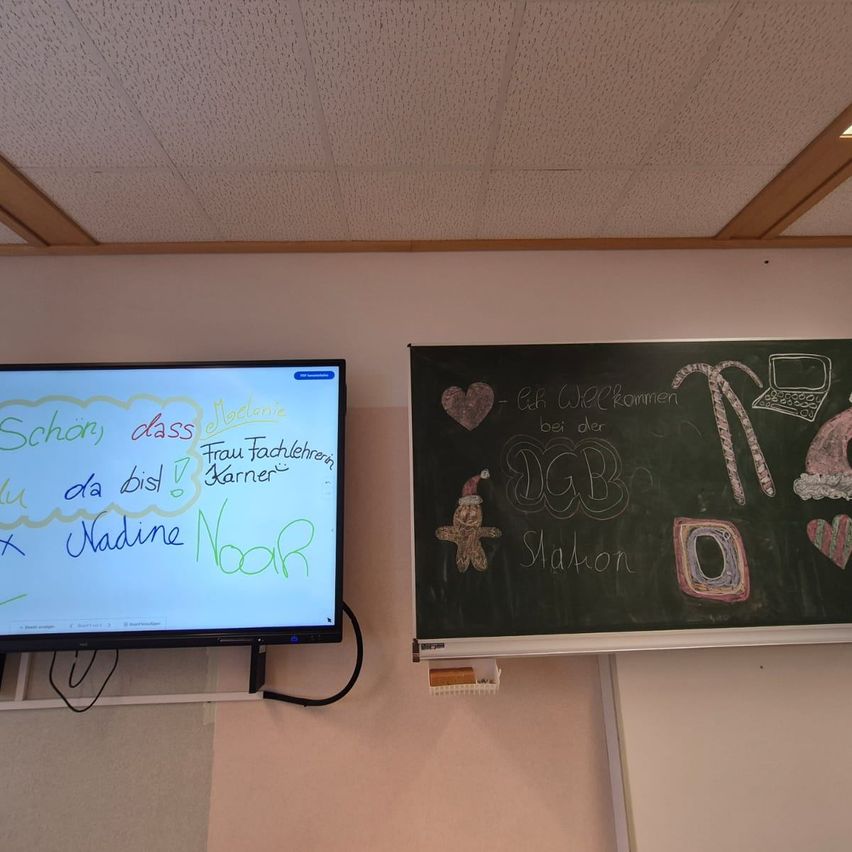 Ein Klassenzimmer mit Tafel und Monitor. Der Monitor zeigt handgeschriebenen Text in mehreren Farben. Die Tafel hat Zeichnungen und Text.