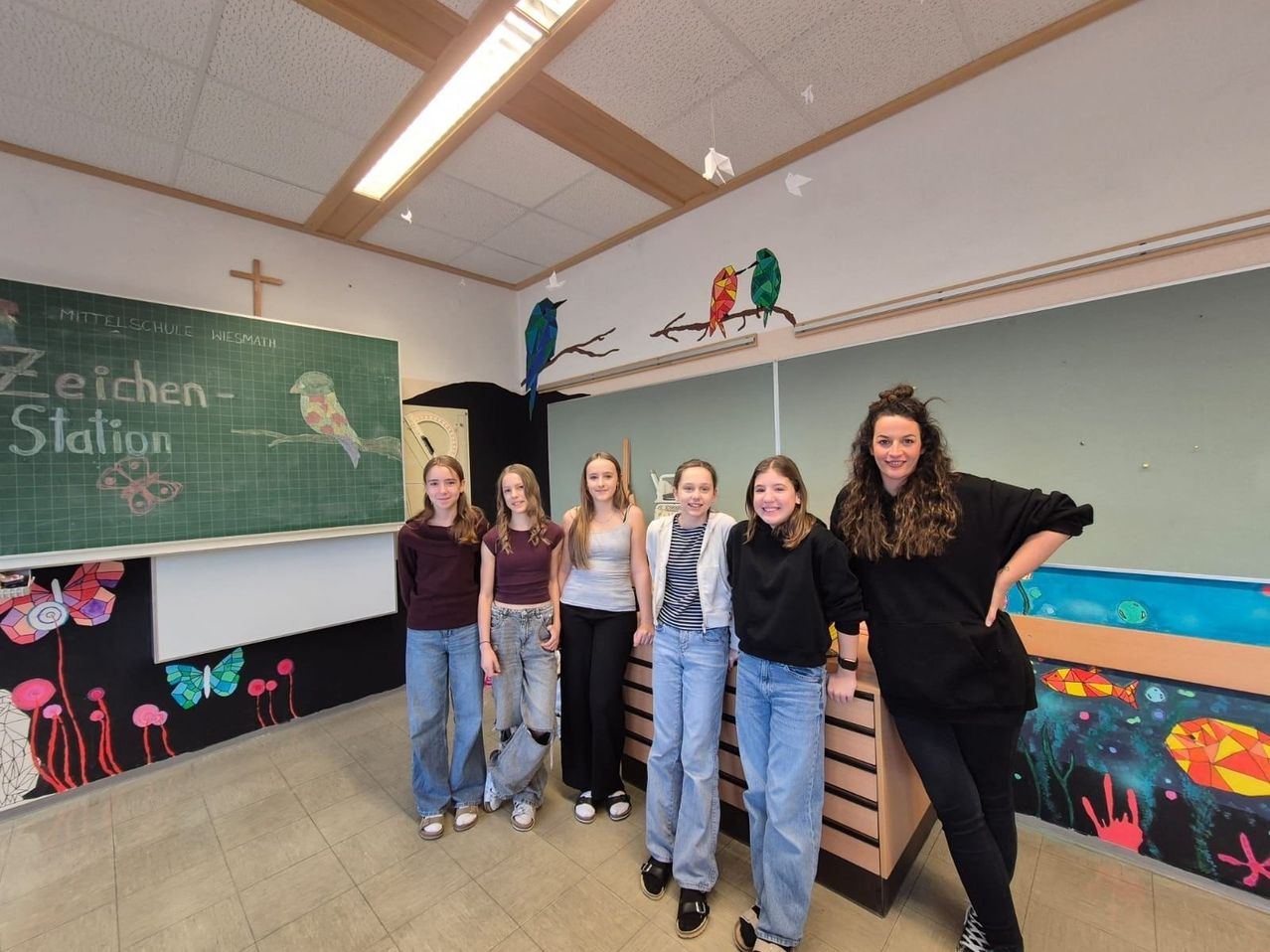Fünf Mädchen und eine Frau stehen in einem Klassenzimmer mit bunten Wanddekorationen und einer Tafel mit Zeichnungen. Sie alle lächeln.