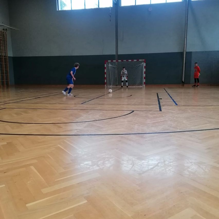 Eine Gruppe von drei Spielern spielt Fußball auf einem Innenplatz. Ein Spieler ist kurz davor, den Ball zu schießen.