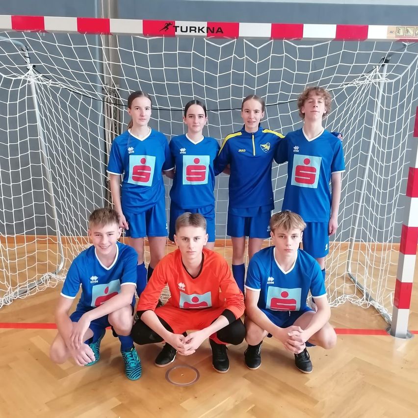 Eine Gruppe junger Athleten in blauen und orangefarbenen Trikots posiert für ein Foto vor einem Fußballtor.