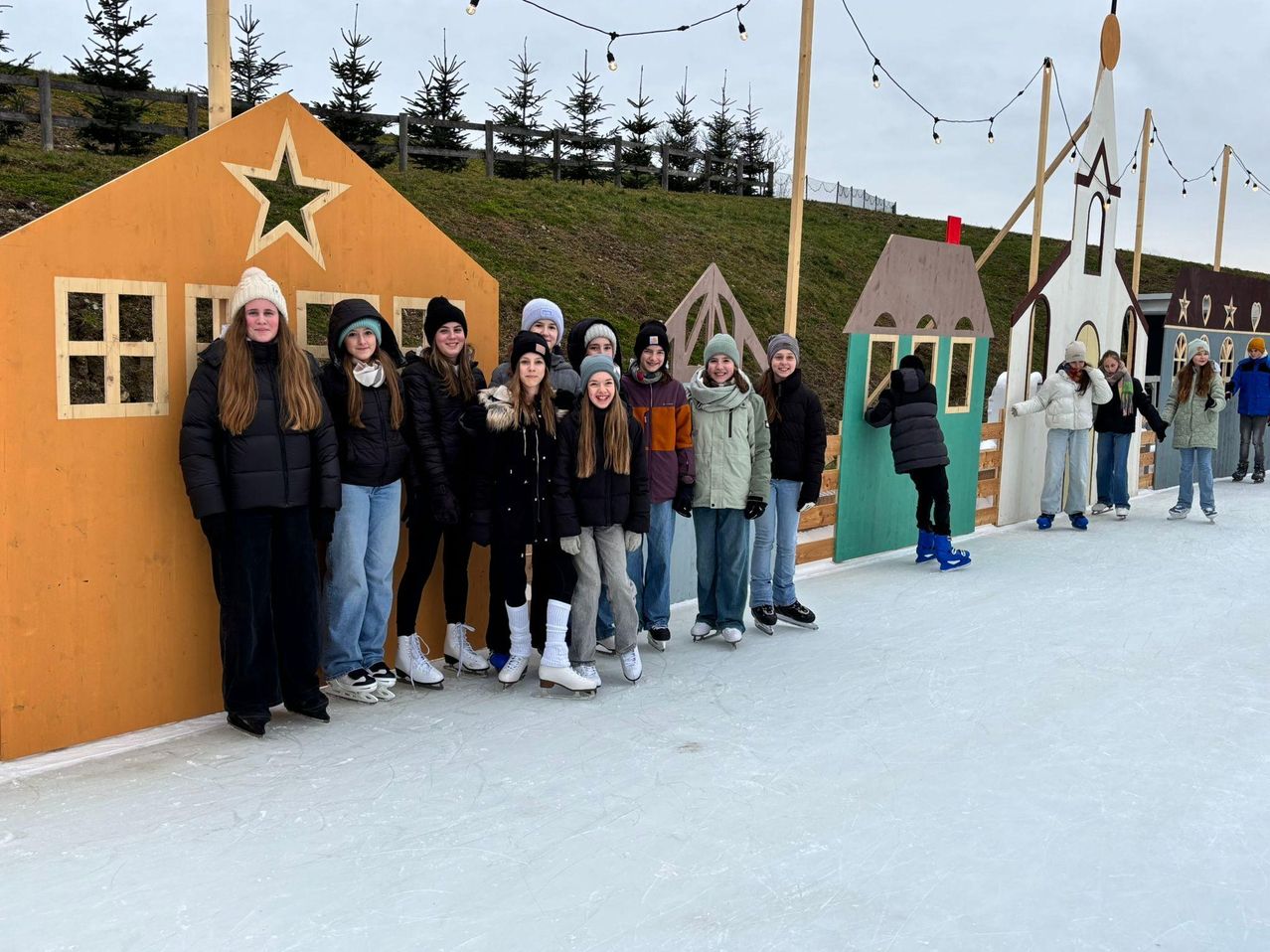 Eine Gruppe von Menschen in Winterkleidung und Schlittschuhen posiert für ein Foto auf einer Eisbahn, mit dekorativen Häusern im Hintergrund.