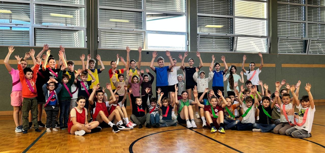 Gruppe von Kindern und Jugendlichen mit erhobenen Händen in einer Turnhalle. Sie tragen verschiedene Farben von Hemden und Hosen, einige haben Medaillen. Der Raum hat einen Holzboden und Glasfenster mit Jalousien.