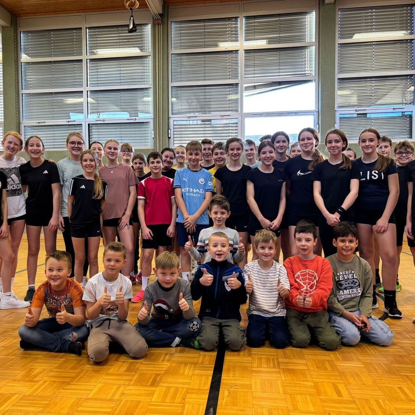 Eine Gruppe von Kindern posiert für ein Foto in einer Turnhalle, einige geben Daumen hoch. Dahinter stehen andere Kinder in zwei Reihen.