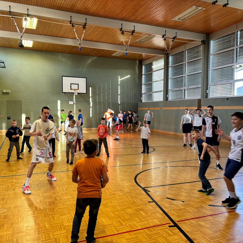 In einer Turnhalle ist eine Gruppe von Kindern in ein lebhaftes Basketballspiel verwickelt. Ein Mann in einem weißen Shirt und Sneakers führt das Spiel an. Andere Kinder spielen in der Nähe, während andere zuschauen und von den Seitenlinien anfeuern.